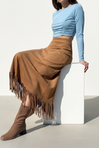 Jupe midi velours camel avec franges - vue assise montrant le drapé naturel de la jupe velours camel en position assise, confort et élégance, franges décoratives marron visibles, texture velours douce, mannequin sur cube blanc en crop top bleu, fond blanc studio BLACK-COAT"