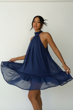 Robe mini halter Black-Coat bleu marine avec double volant - vue trois-quarts d'une mannequin en léger mouvement de rotation sur fond blanc studio, capturant la richesse de la couleur bleu marine profond, le mouvement fluide des deux étages de volants, la coupe halter élégante et la silhouette féminine moderne
