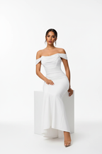 "Robe sirène maxi blanche épaules dénudées froncée - vue assise montrant le drapé naturel de la robe en position assise, confort et élégance, décolleté bardot avec manches drapées, silhouette moulante, mannequin sur cube blanc, fond blanc studio BLACK-COAT"